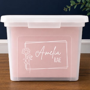 May include: A rectangular, translucent plastic storage bin with a matching lid. The front features a white graphic design consisting of a rectangular frame, a line-art flower, and the personalized text Amelia RAE printed in a white script and sans-serif font.