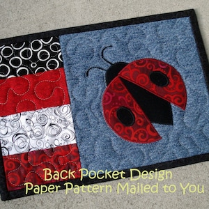 May include: A quilted denim placemat with a ladybug design. The ladybug has red wings with black spots and a black body. The left side features three fabric panels in black and white circles, red swirls, and white swirls. The text "Back Pocket Design Paper Pattern Mailed to You" is at the bottom.
