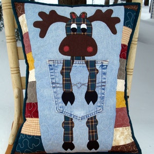 May include: A decorative pillow featuring a moose design. The moose has brown antlers, a brown face, and red nose details. The pillow has a denim pocket and plaid suspenders. The pillow is on a wooden chair.