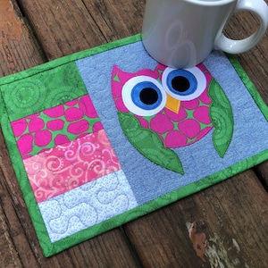 May include: A handmade, square fabric coaster featuring a whimsical owl design. The owl has pink and green patterned feathers, large blue eyes, and a yellow beak. The coaster has a denim blue background and a green border. A white mug rests on the coaster.