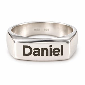 May include: A polished silver signet ring featuring a flat rectangular face engraved with the name Daniel in a bold black font. The interior of the band is stamped with the text MEX 925, indicating the metal composition and origin.