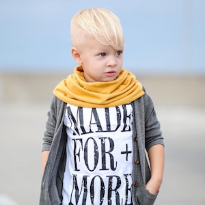 May include: A young boy wearing a gray cardigan, a white t-shirt with the text "MADE FOR + MORE" printed on it, and a yellow scarf.