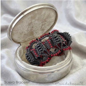 May include: A beaded bracelet with black and red beads rests inside a silver jewelry box. The bracelet features a repeating pattern of black oval beads and small red beads. The jewelry box is open, revealing a soft, light-colored interior.
