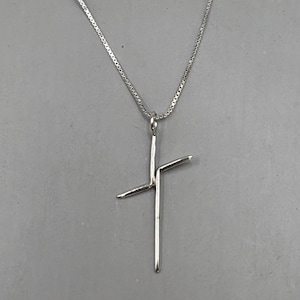 May include: A silver-colored pendant necklace featuring a cross design. The cross has a vertical bar with a horizontal bar extending from the center. The necklace chain is a delicate, box-style chain.
