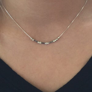 May include: A delicate silver necklace with a series of small, sparkling beads and darker, cylindrical beads. The necklace is worn against a person's skin, with a black top visible in the background. The necklace is a simple, elegant piece of jewelry.