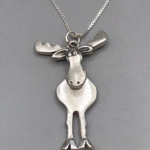 May include: A silver moose pendant necklace with a detailed design. The moose has large antlers, a round body, and small legs. The necklace is on a silver chain.