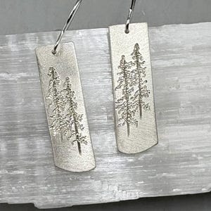 May include: A pair of silver earrings with a rectangular design featuring a detailed etched image of two pine trees.