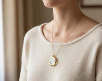 May include: A gold-colored locket necklace with a round pendant. The pendant has a clear front, revealing a white interior. The necklace is worn against a cream-colored sweater.