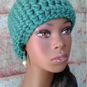 May include: A teal crochet hat with a textured stitch pattern. The hat is worn by a mannequin with dark hair and a pair of dangling earrings with pink, yellow, and blue beads. The background is a rustic, multi-colored wall.