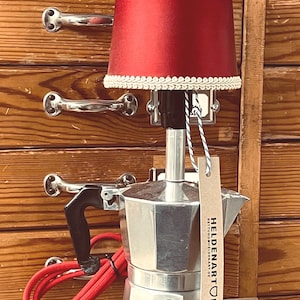 Puede incluir: Una lámpara única hecha de una cafetera espresso plateada, coronada con una pantalla roja adornada con encaje blanco. La lámpara tiene un asa negra y un cable rojo. Una etiqueta con el texto "HELDENART.DE" cuelga de la lámpara.
