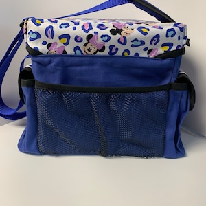 Puede incluir: Una bolsa de lona azul y blanca con estampado de Minnie Mouse. La bolsa tiene una parte superior con cremallera, un bolsillo de malla y una correa para el hombro.