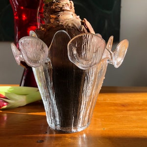 May include: A clear glass vase with a textured surface and a unique, organic shape. The vase has decorative, petal-like elements around the rim. The vase is sitting on a wooden surface, with a red vase and plant in the background.