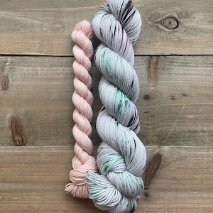 May include: Two skeins of yarn are displayed against a wooden background. One skein is a solid light pink, while the other is a variegated yarn with shades of gray, green, and black. The yarn is tightly wound, showcasing its texture and color variations.