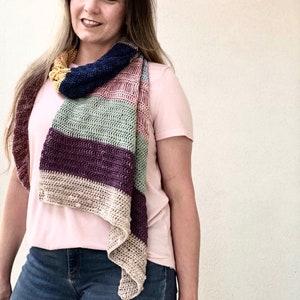 May include: A woman wearing a pink t-shirt and blue jeans is wearing a multi-colored crocheted scarf. The scarf has stripes of yellow, blue, pink, green, purple, and beige.
