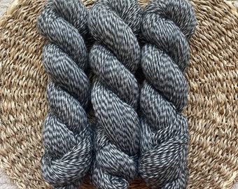 Puede incluir: Tres madejas de hilo moteado gris y blanco están dispuestas sobre una cesta redonda de mimbre. El hilo está bien enrollado, mostrando una superficie texturizada. La cesta está hecha de fibras naturales, y el fondo es una alfombra clara y texturizada.