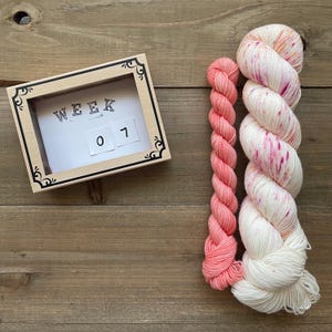 Puede incluir: Un calendario con marco de madera muestra "WEEK 07" junto a dos madejas de hilo. Una madeja es un degradado de rosa coral, y la otra es blanca con motas rosas. Los artículos están sobre una superficie de madera.