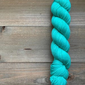 May include: A skein of vibrant turquoise yarn, tightly wound and displayed against a rustic wooden background. The yarn's texture is clearly visible, showcasing its fine strands and potential for knitting or crocheting projects. The color is a bright, solid turquoise.