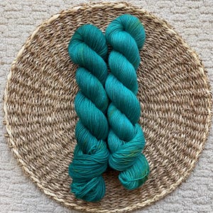 May include: Two skeins of teal-colored yarn are displayed on a round woven mat. The yarn has a slight sheen and a variegated color pattern with hints of yellow and green. The mat is made of natural fibers.