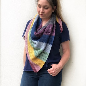 Op de afbeelding: Een vrouw die een blauw shirt en jeans draagt, draagt een kleurrijke gehaakte sjaal met verschillende kleuren, waaronder geel, groen, blauw en paars.