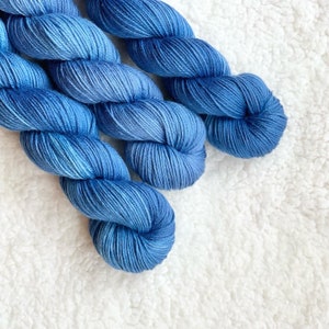 May include: Close-up of three skeins of blue yarn with varying shades of blue. The yarn is tightly wound and has a soft, textured appearance. The background is a white, fluffy surface. Ideal for knitting and crochet projects.