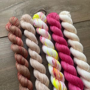 May include: Five skeins of colorful yarn in a row. The yarn colors include shades of brown, tan, yellow, pink, and white. The yarn is twisted into a rope-like shape, and the background is a wooden surface.