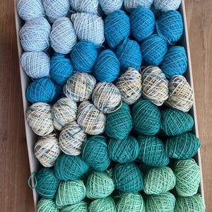 May include: A rectangular box filled with numerous small, round balls of yarn arranged in a color gradient. The collection transitions from light sky blue and turquoise to deep teal, forest green, and pale mint, with some variegated white and yellow strands.