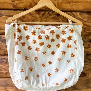 May include: A white fabric square featuring a repeating pattern of small, five-petaled orange flowers. The textile is suspended from a wooden hanger, displaying a clean, rectangular shape with visible stitching along the bottom edge and sides.