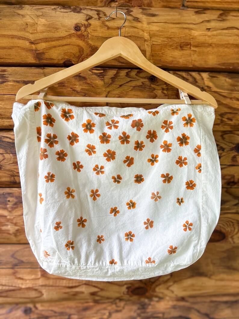 May include: A white fabric square featuring a repeating pattern of small, five-petaled orange flowers. The textile is suspended from a wooden hanger, displaying a clean, rectangular shape with visible stitching along the bottom edge and sides.