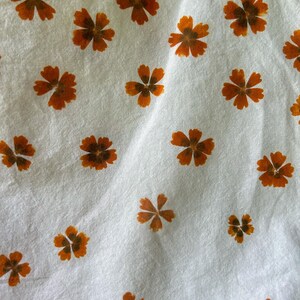 May include: A white fabric featuring a repeating pattern of small, five-petaled orange flowers. The flowers are distributed across the surface in a scattered arrangement, each displaying a darker brown center and distinct petal shapes.
