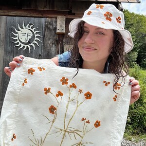 May include: A white bucket hat and a matching white tote bag, both decorated with a botanical print of orange flowers and green stems. The fabric items feature a repeating pattern of small floral motifs, with the tote bag displaying a larger central arrangement of stems and blossoms.