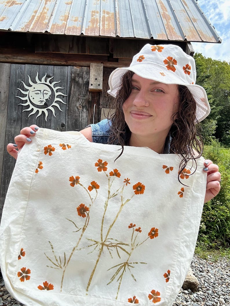 May include: A white bucket hat and a matching white tote bag, both decorated with a botanical print of orange flowers and green stems. The fabric items feature a repeating pattern of small floral motifs, with the tote bag displaying a larger central arrangement of stems and blossoms.