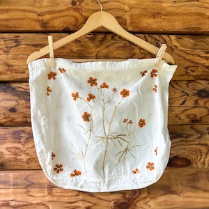 May include: A white fabric tote bag featuring a botanical print of orange flowers with green stems and leaves. The bag is suspended from a light-colored wooden hanger by two wooden clips, displaying a flat, rectangular shape with a simple design.