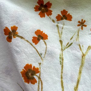 May include: A white fabric featuring a botanical print of orange flowers with dark brown centers and thin, textured green stems. The floral pattern is scattered across the textile, showcasing delicate petals and slender stalks in a natural arrangement.