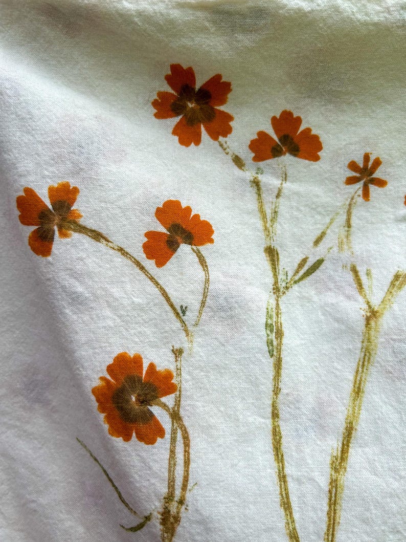 May include: A white fabric featuring a botanical print of orange flowers with dark brown centers and thin, textured green stems. The floral pattern is scattered across the textile, showcasing delicate petals and slender stalks in a natural arrangement.