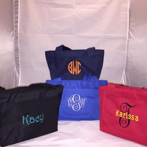 May include: Four different tote bags with personalized embroidery. The bags are navy blue, black, blue, and red. The embroidery includes the names "Kacy", "BWC", "LES", and "Karissa".