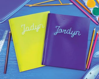 May include: Two personalized notebooks, one yellow and one purple, are displayed on a blue surface. The name "Jadyn" is written in white script on the yellow notebook, and "Jordyn" is written in blue script on the purple notebook. Pencils, a ruler, and paint are also visible.