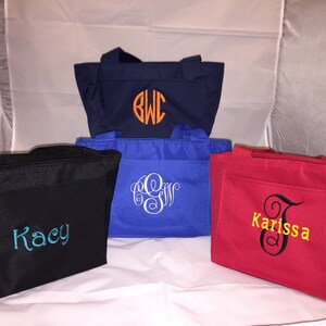 May include: Five different tote bags with personalized embroidery. The bags are in various colors, including black, navy blue, red, and blue. The embroidery includes names and initials.