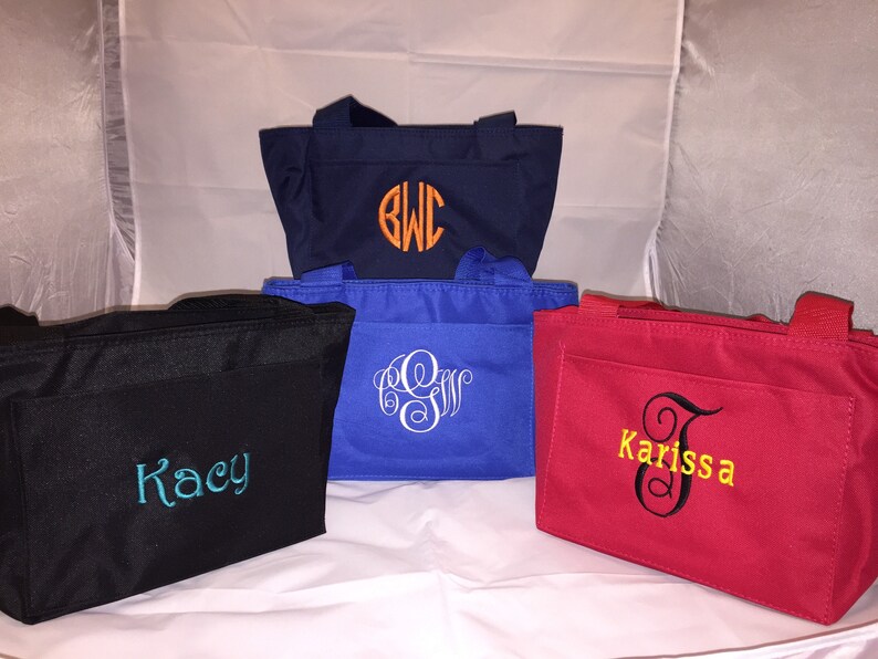 May include: Five different tote bags with personalized embroidery. The bags are in various colors, including black, navy blue, red, and blue. The embroidery includes names and initials.