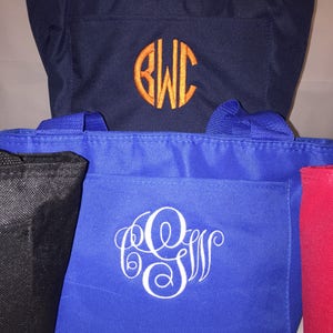 May include: Three tote bags with different colors and monograms. The top bag is navy blue with an orange monogram "BWC". The middle bag is royal blue with a white monogram "GSW". The bottom bag is black.