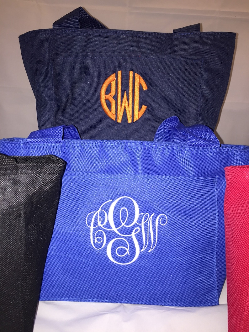 May include: Three tote bags with different colors and monograms. The top bag is navy blue with an orange monogram "BWC". The middle bag is royal blue with a white monogram "GSW". The bottom bag is black.