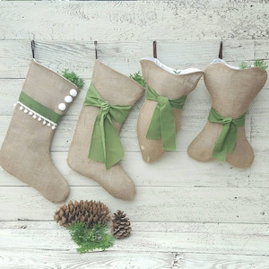 May include: Four holiday stockings made of tan hessian fabric. Two are traditional stocking shapes, one with white pom-pom trim and a green ribbon, the other with a green bow. Two are bone-shaped with green ribbons. Pinecones and greenery are visible.
