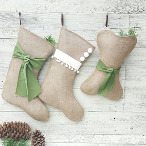 Peut inclure: Trois chaussettes en toile de jute suspendues à des crochets. Deux chaussettes ont une forme traditionnelle avec des nœuds en tissu vert, tandis que celle du milieu est ornée d'un ruban blanc et de pompons blancs. Chaque chaussette contient des brins de pin.