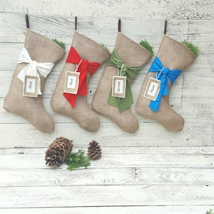 May include: Four hessian Christmas stockings with hanging loops, each featuring a large fabric bow in white, red, olive green, or blue. Each stocking includes a rectangular tag with the printed names Mom, Dad, Garrett, and Ryan respectively.