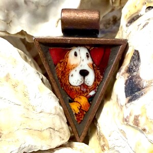 May include: A triangular pendant featuring a copper-toned metal frame and a central graphic illustration of a brown and white dog with floppy ears. The dog is depicted against a red background, and the pendant includes a top metal loop.
