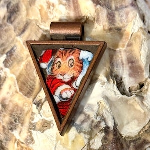 May include: A triangular pendant with a copper-toned metal frame featuring an illustration of an orange tabby cat wearing a red and white Santa hat. The cat is depicted with large eyes and a white paw reaching forward against a blue background.