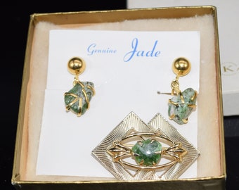Può includere: Un set di gioielli in giada in una scatola. Il set include una spilla con una pietra di giada verde in una montatura dorata e un paio di orecchini con pietre di giada verdi.