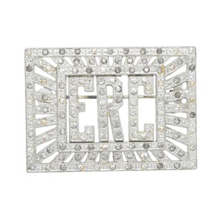 May include: A rectangular silver-toned brooch with the letters "ERC" in the center. The brooch is encrusted with small, sparkling stones, and the frame has a geometric design with radiating lines.
