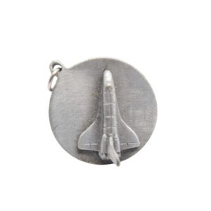 May include: A circular silver-toned metal charm featuring a raised, three-dimensional space shuttle design centered on the disc. The charm includes a small metal jump ring attached to the top edge for hanging, with a matte, brushed finish.