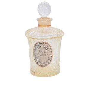 Pu&ograve; includere: Un'antica bottiglia di profumo con un corpo in vetro trasparente e scanalato e un tappo decorativo trasparente. La bottiglia presenta un'etichetta con il testo "GUERLAIN Apr&egrave;s l'Ond&eacute;e". La bottiglia ha un'estetica vintage.