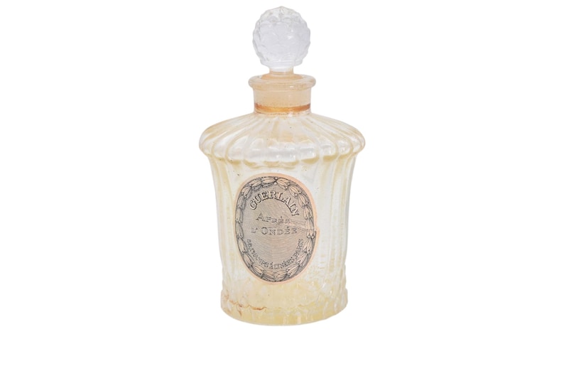 Pu&ograve; includere: Un'antica bottiglia di profumo con un corpo in vetro trasparente e scanalato e un tappo decorativo trasparente. La bottiglia presenta un'etichetta con il testo "GUERLAIN Apr&egrave;s l'Ond&eacute;e". La bottiglia ha un'estetica vintage.